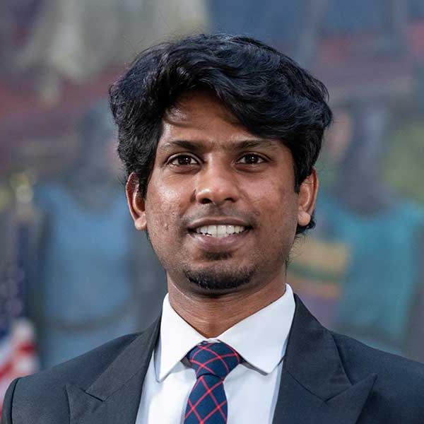 A man with tanned skin and black hair poses. He wears a black dress suit, a white dress shirt, and a blue-and-red tie. Blurred in the background is an American flag and a mural.